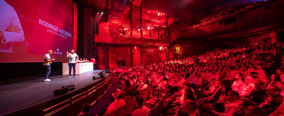 conference hall stage with two speakers presenting to a large red lit audience on a packed venue  youprotv