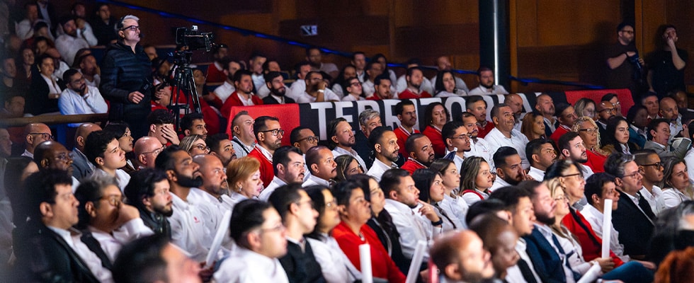 large audience seated in a dim auditorium attentive to a stage with a videographer on a tripod at the back left  youprotv
